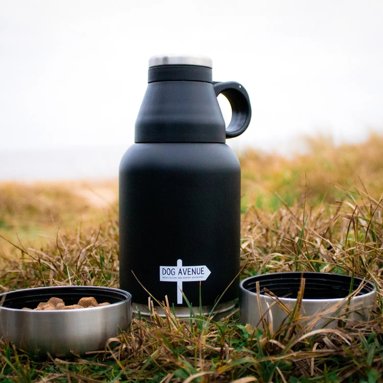 Matte black 3-in-1 dog water bottle standing in a grassy field flanked by its two detachable bowls, one filled with treats